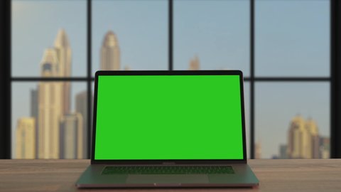 Modern notebook with open green mock-up template monitor on office desk close-up. Working space with laptop computer on Dubai cityscape background.