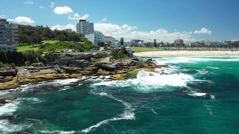 Sydney, Australia. Bondi beach, reveal drone video clip. Beautiful sunny day, big Pacific Ocean waves hitting the rocks. Stunning clear blue ocean water. Most popular white sand beach in Sydney.