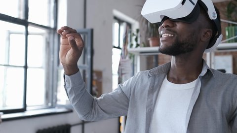 Handsome african american man wearing virtual reality headset. Augmented Reality. Man touch something using modern 3D vr glasses indoors. Student playing using VR glasses on a sunny day. Shot on 4K