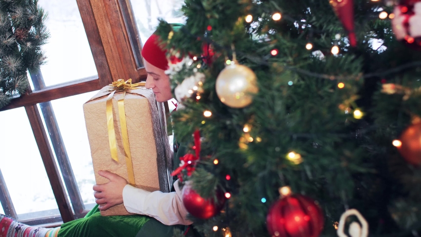 Elf yawns and sits on a windowsill near a Christmas tree with decorations hugging a box with gifts