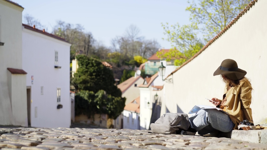 Young woman walking at downtown of Prague. Elegant lady sitting on cobblestone pathway between old houses. Girl wearing coat and hat checking the map and enjoying sunny day at narrow city street
