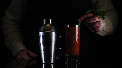 Bartender mixes a Bloody Mary drink cocktail in a shaker and pours into a glass with ice cubes and a celery against a black background