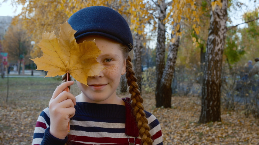 Portrait teenager girl with yellow maple leaf in hand near face. Smiling face happy girl in autumn park. Cheerful red haired girl looking to camera in yellow city park