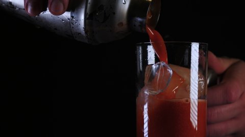 Bartender mixes a Bloody mary drink and pours into a glass filled with ice and garnishes with a celery stick against black background in close up