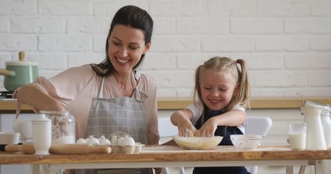 Cute small kid daughter learning kneading dough with hands helping mom in modern kitchen, happy family adult mother and little child girl prepare cookies biscuit having fun cooking together at home