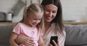 Happy family mom embrace cute little kid child daughter sit on sofa laugh having fun holding using smart phone watching funny social media video app, take selfie looking at cellphone screen at home