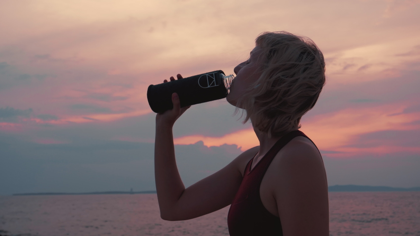 Close up shot of a female who finished her run, drinking the water from her reusable glass bottle and the sunset/sunrise. Zero waste lifestyle.