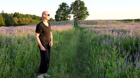A young bald man black casual clothes, black sunglasses, mirrored glasses, jumps and waves his legs depicting karate fighting techniques. In summer, summer green flowering field. Slow motion video.
