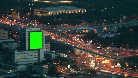 Large empty billboard with a green screen for advertising on the modern building near speed highway, urban interchange top view, timelapse of traffic at night 