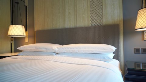 Sliding shot of a hotel room bed covered in white and blue linens with a big wooden headboard and sidelamps