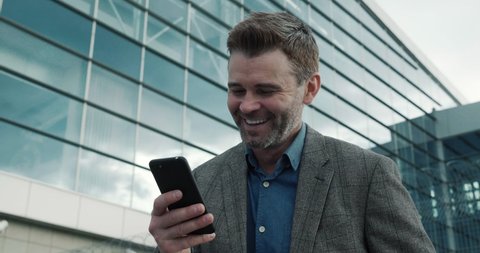 Closeup of satisfied Man browsing his Smartphone, glassy Building on Background. Bearded Businessman smiling cute while texting Messages and browsing Social Network. Emotions. People. Phones. Apps.