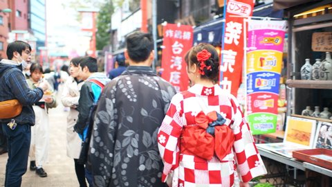 Tokyo, Japan - March 30, 2018: Panning, point of view walking pov shot of Asakusa with young Chinese tourists couple dressed up in red Kimono costumes back on Nakamise street by food vendors