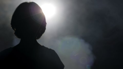 dark silhouette of girl with short hair mimes raising hand with open palm to bright light in clouds of smoke slow motion