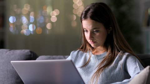 Sad woman reading bad news on laptop sitting on a couch in the night at home