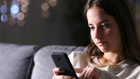 Sad woman reading mobile phone content sitting on a couch at home in the night