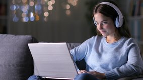 Concentrated woman wearing headphones writing on laptop sitting on a couch in the night at home