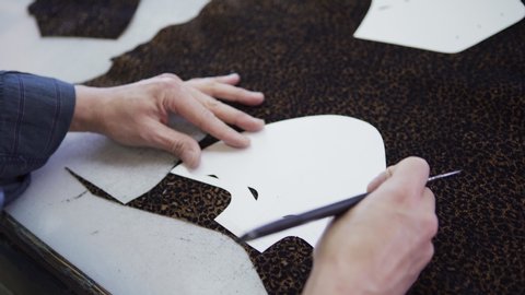 Hands of skilled shoemaker using paper pattern and leather knife to cut out shoe part from piece of leopard print textile standing at work table, high angle view closeup shot