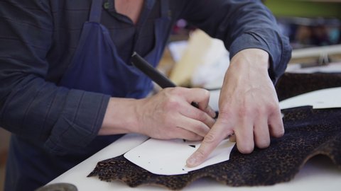 Side view of unrecognizable shoemaker using paper patterns and leather knife to cut out shoe parts from piece of leopard print textile standing at workbench in studio, closeup