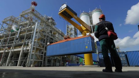 yellow crane with large blue container and worker with remote control at gas oil refinery plant on sunny day backside view