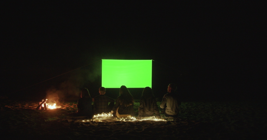 Outdoor cinema. Friends gather in a campfire camp and watch a movie with a projector on the beach near the sea. Green screen. A place for your image.