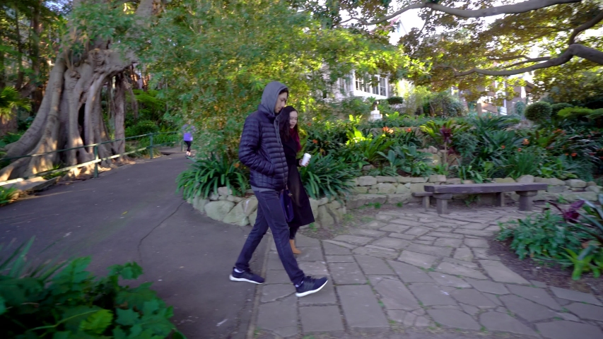 Couple of Travelers Walking and Talking in Public Park. Female in Coat With Coffee To Go and Male in Hoodie