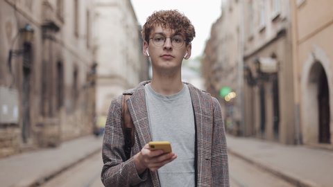 Close Up of Calm Millenial Hipster Guy in Earphones and Glasses Looking to Camera Holding Smartphone in his Hand Standing at Blured Old City Background Zoom.