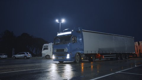 Blue Semi-Truck with Cargo Trailer Drives Into Overnight Parking Space where Other Trucks are Standing. Drivers Resting at Night on the Overnight Parking Lot
