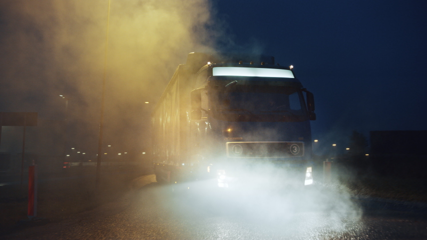 Blue Long Haul Semi-Truck with Cargo Trailer Full of Goods Travels At Night , Turning on the Freeway Road, Driving Across Continent Through Rain, Fog, Snow. Industrial Warehouses Area. Front Following Royalty-Free Stock Footage #1039037177