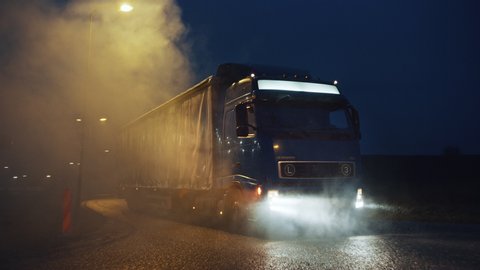 Blue Long Haul Semi-Truck with Cargo Trailer Full of Goods Travels At Night , Turning on the Freeway Road, Driving Across Continent Through Rain, Fog, Snow. Industrial Warehouses Area. Front Following