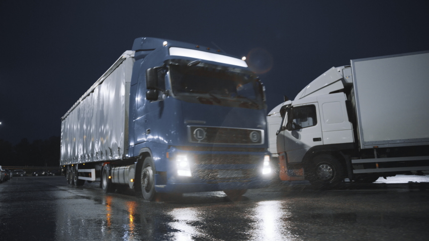 Blue Semi-Truck with Cargo Trailer Drives Off From Overnight Parking Space where Other Trucks are Standing. Long Haul Truck Leaves Parking Lot, Transporting Cargo / Goods Across Continent. Rainy Night Royalty-Free Stock Footage #1039037180