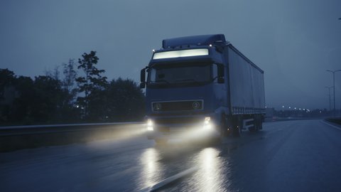 Long Haul Semi-Truck with Cargo Trailer Full of Goods Travels on the Highway Road. Driving in Early Morning Across Continent Through Rain, Fog. Industrial Warehouses Area. Front Overtaking Camera Shot