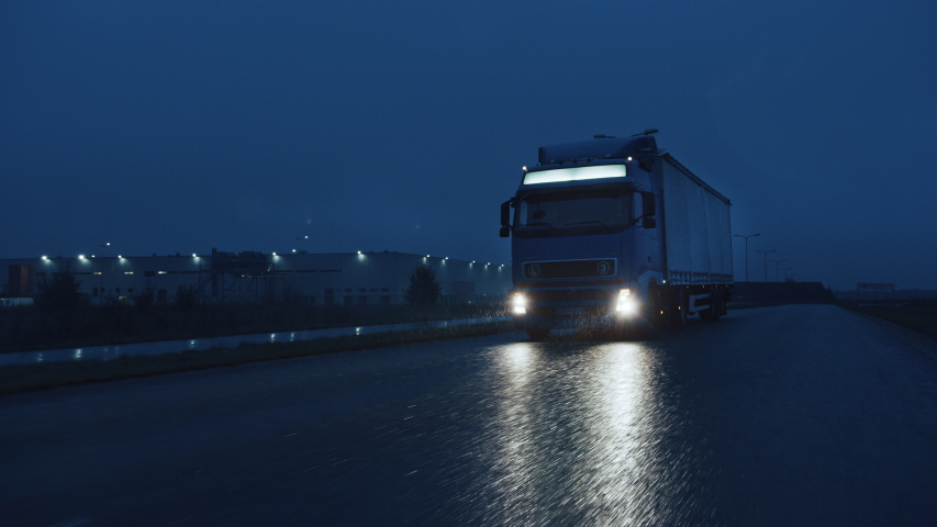 Long Haul Semi-Truck with Cargo Trailer Full of Goods Travels At Night on the Freeway Road, Driving Across Continent Through Rain, Fog, Snow. Industrial Warehouses Area. Front Following Slow Motion  Royalty-Free Stock Footage #1039037240