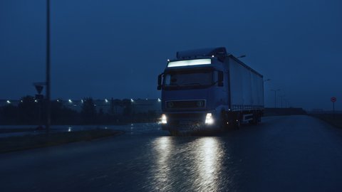 Long Haul Semi-Truck with Cargo Trailer Full of Goods Travels At Night on the Freeway Road, Driving Across Continent Through Rain, Fog, Snow. Industrial Warehouses Area. Front Following Slow Motion 