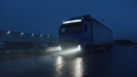 Blue Long Haul Semi-Truck with Cargo Trailer Full of Goods Travels At Night on the Freeway Road, Driving Across Continent Through Rain, Fog, Snow. Industrial Warehouses Area. Front Following Shot
