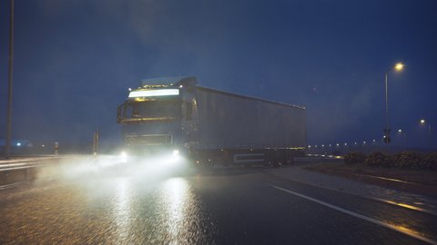 Blue Long Haul Semi-Truck with Cargo Trailer Full of Goods Travels At Night on the Freeway Road, Driving Across Continent Through Rain, Fog, Snow. Industrial Warehouses Area. Front Following Shot