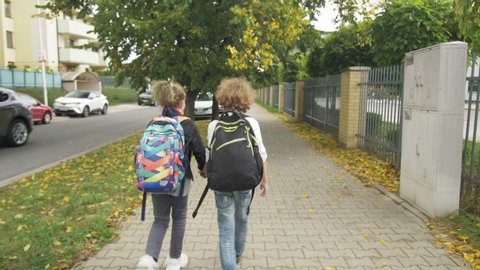 Two Friends with Backpacks are Going to School on City Streets past Cars. Sister with her Brother Back to School. Sun is on Background.