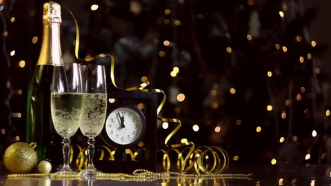 Glasses with champagne, new year golden decor, balls are on table. Black clock is ticking, few minutes to midnight. Festive decorative garland, warm yellow light bulbs are blinking on background.