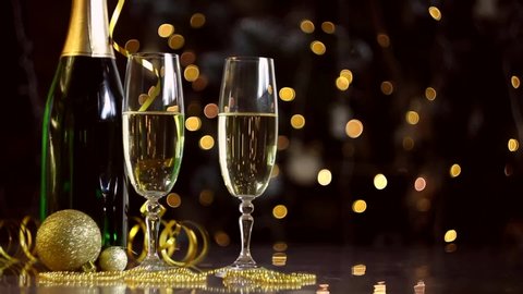Female hand is taking glass with champagne. Golden new year christmas decor, balls, green bottle are on table. Festive decorative garland, warm yellow light bulbs are blinking on background.
