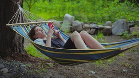 Relaxed young beautiful girl lying in a hammock on vacation. A woman scrolls through photos on the phone, photographs herself on a smartwatch. 4k video.