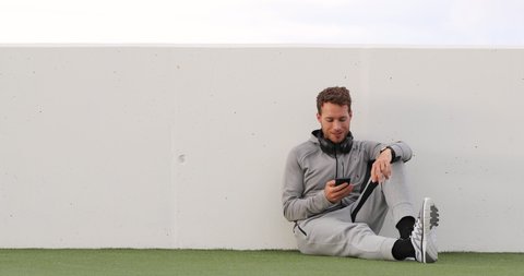 Man using phone looking at smartphone app sitting outdoors wearing headphones sitting outdoors on grass. Healthy lifestyle sport athlete using smartphone on jogging break. 59.94 FPS slow motion.