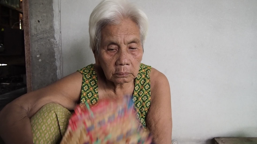 Aged senior woman or old grandmother with white grey hair holding hand fan and blowing for wind at home. Lifestyle retirement time in rural countryside.