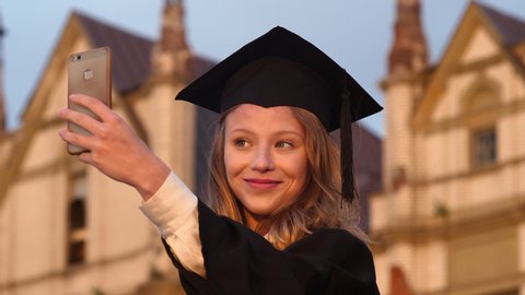 Attractive graduate girl taking selfie on graduation day holding diploma.
