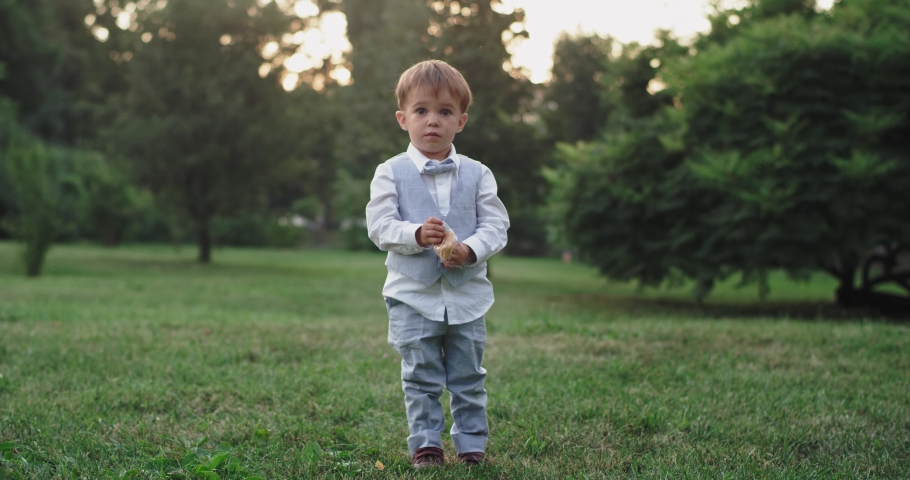 Lovey little boy eating his snacks in front of the camera in the middle of a green field in the central park wearing a cute casual suit
