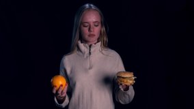 Pretty blondie woman in white sweater choosing between orange and burger over black background. Healthy food. Girl on a diet.