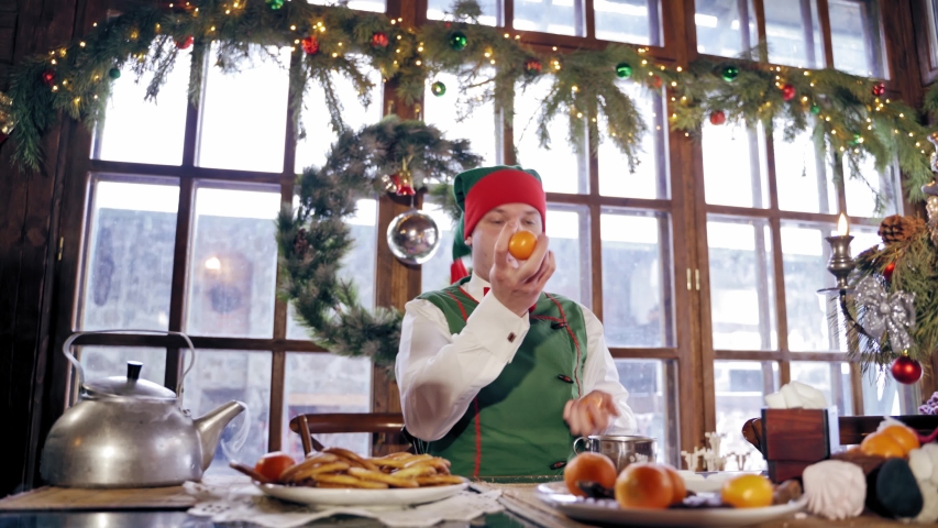 Elf playing tangerines at a table. Santas sitting at Christmas table in a decorated room. New Year celebration. Christmas eve.