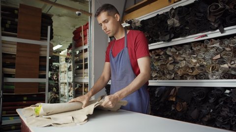 Side view of young shoemaker in apron unrolling and checking pieces of leather standing at table in material storage