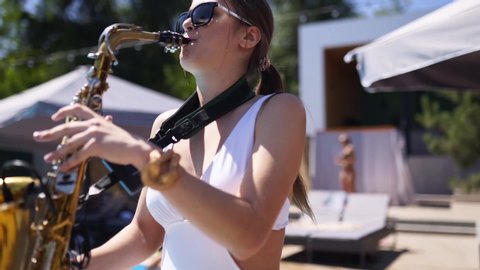 Attractive saxophonist lady is playing on saxophone near pool at beach club. Pretty sax girl musician in hot white bikini dances and plays on weekend party onhot summer day.