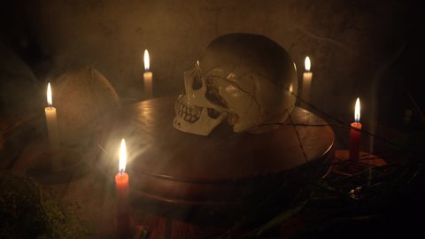 Human skull rotating on a wooden circular platform with dark gray background with candles and mystic herbs around a table with smoke