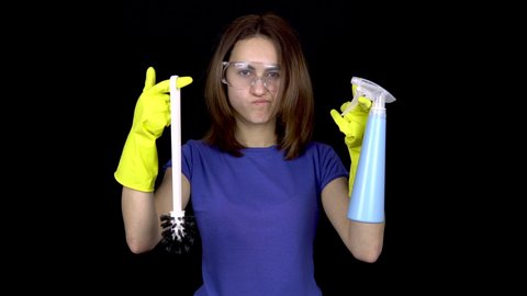 A young woman does not want to do the toilet cleaning. Woman in safety glasses and gloves with tools for cleaning the toilet. Girl holds a toilet brush and water spray. On a black background
