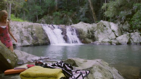 Two beautiful young women undress to bikinis as they are about to swim in a clear pool in a stream with rocky banks with a waterfall in the background and a with towels and a camera in the foreground,